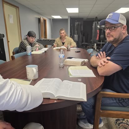 Several men gathered around a wood conference table for Bible study in a room with gym and boxing equipment.