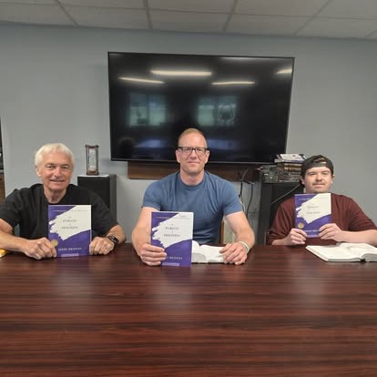 Three men seated at a table holding copies of a book during a study session in front of a TV.