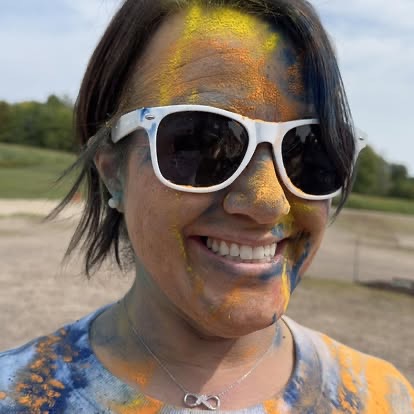 A woman smiling brightly, her face and sunglasses covered in blue and orange powder at a community color run.