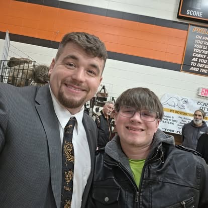 A man in a suit taking a friendly selfie with a teenager at a local school gym community event.