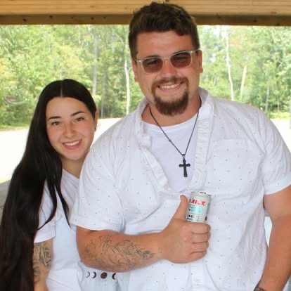 A man and woman smiling together under a wooden park pavilion at a summer community gathering.