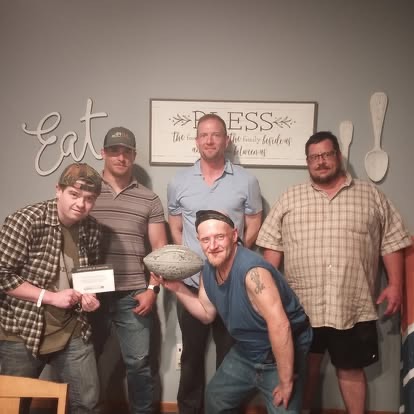 Five men posing together indoors beneath Eat and Bless wall decor, one holding a football.
