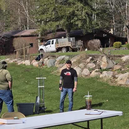 Two residents setting up folding tables and gear on the lawn in front of the lodge and a pickup truck before an outdoor event.