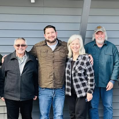 Four people standing arm in arm in front of a gray house exterior, residents, family and supporters together.