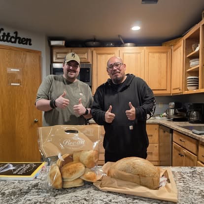 Two men giving a thumbs up together in the His House of Hope kitchen with fresh bread on the counter