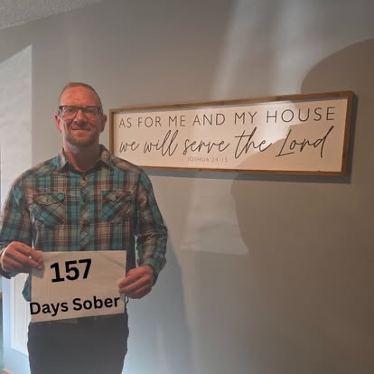 A man in a plaid flannel shirt holding a 157 Days Sober sign beneath the As For Me And My House Scripture sign.