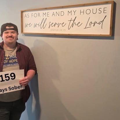 A young man holding a 159 Days Sober sign beneath the As For Me And My House We Will Serve The Lord scripture sign.