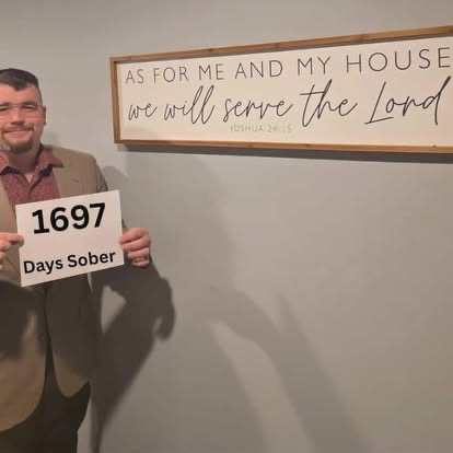 A man in a tan blazer holding a sign reading 1697 Days Sober beneath the As For Me And My House Scripture sign.