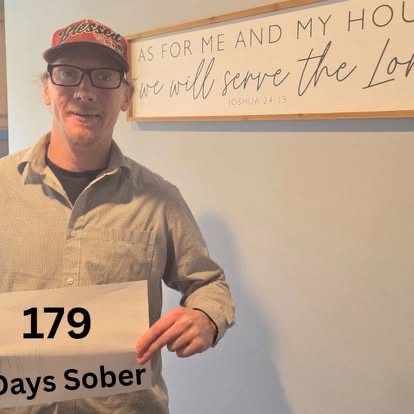A man in a tan button-up shirt holding a 179 Days Sober sign beneath the As For Me And My House Scripture sign.