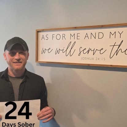 A man in a cap holding a 24 Days Sober sign beneath the As For Me And My House Scripture sign.