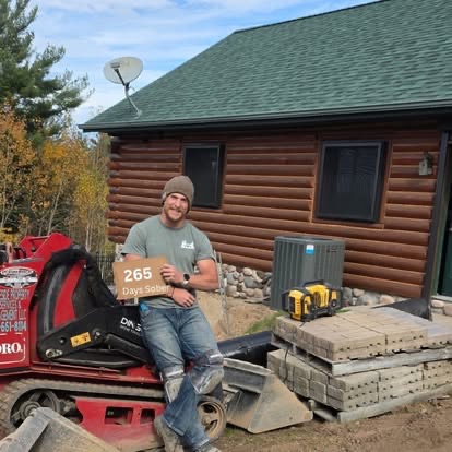 A man sitting on a red skid-steer loader in front of a log cabin holding a 265 Days Sober sign during autumn landscaping work.