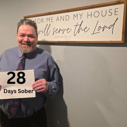 A smiling man in a shirt and tie holding a 28 Days Sober sign beneath the As For Me And My House Scripture sign.