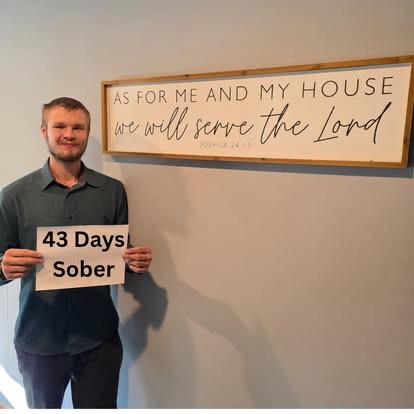 A man holding a 43 Days Sober sign beneath the As For Me And My House We Will Serve The Lord scripture sign.