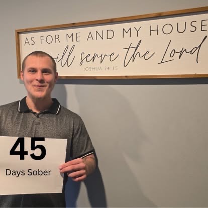A young man in a gray polo holding a 45 Days Sober sign beneath the As For Me And My House We Will Serve The Lord Scripture sign.