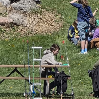 A resident seated at a picnic table by the pond with fishing rods lined up and children playing on the grass nearby.