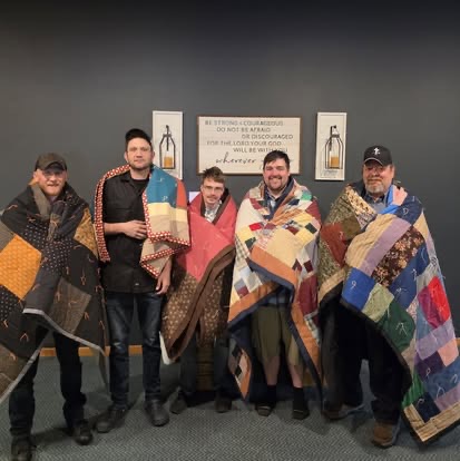 Five men wrapped in handmade quilts standing before a Be Strong And Courageous Scripture sign.