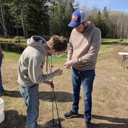 Two young men rigging fishing rods together beside the pond, with forest and grass in the background.