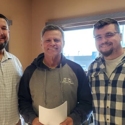 Three men smiling together indoors, the man in the center holding a certificate, at His House of Hope.