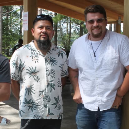 Two men smiling together under a wooden park pavilion at a summer community gathering.